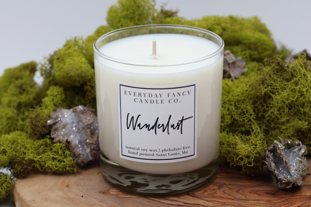 A soy candle labeled 'Wanderlust,' evoking the scent of a misty forest. It features top notes of ozone and bergamot, with calming base notes of moss, juniper, and mint. The candle offers a refreshing, nature-inspired fragrance, perfect for year-round use